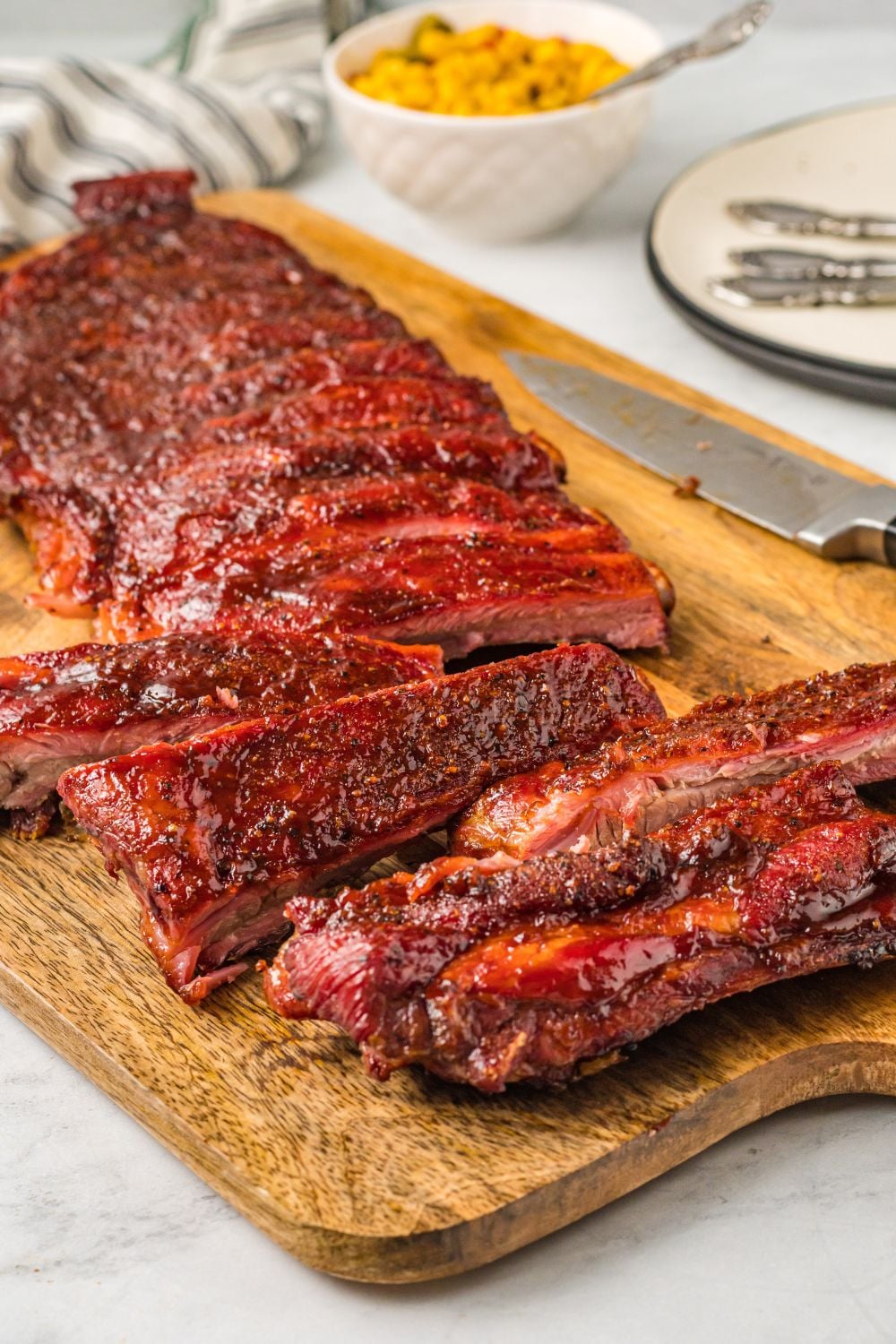 Grilled BBQ Baby Back Ribs sliced up on a cutting board with a bowl of corn in background