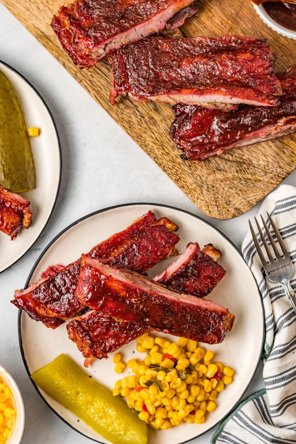 looking down at table with Grilled BBQ Baby Back Ribs on plates and a cutting board with corn and pickles on plates