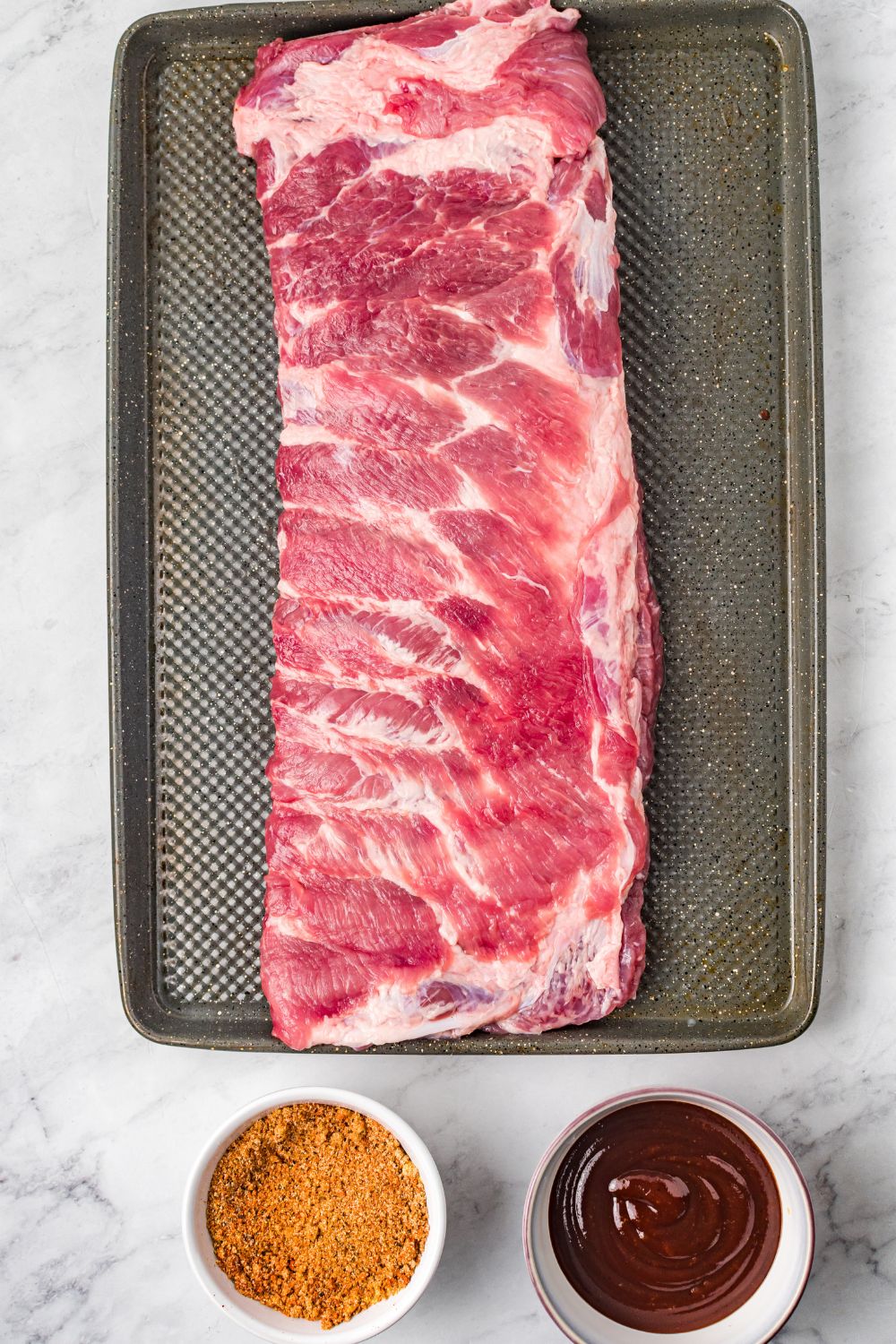 raw ribs on a cooking tray with bbq sauce and dry rub in a bowl