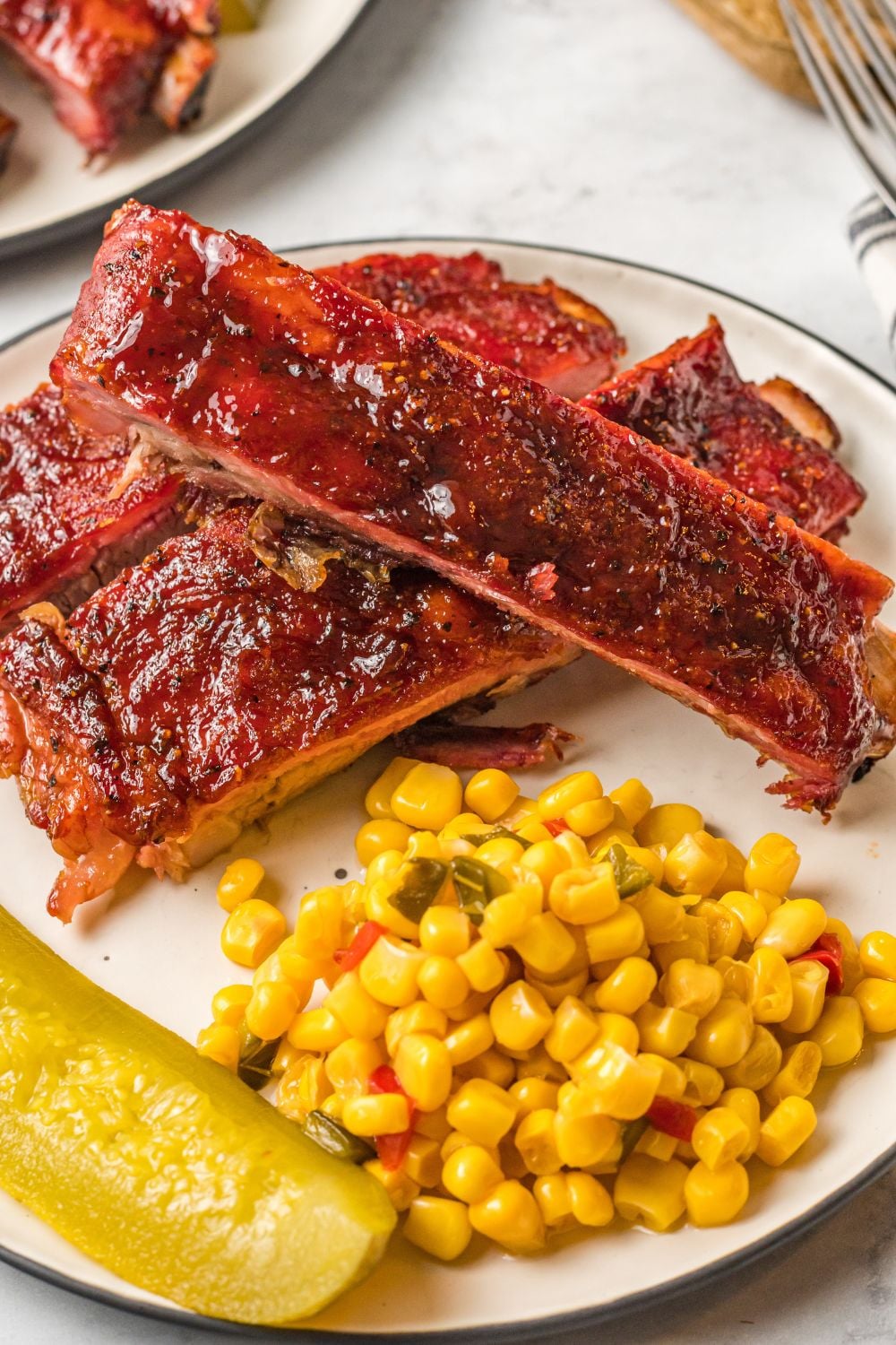 Grilled BBQ Baby Back Ribs on plate with corn and a pickle wedge to side