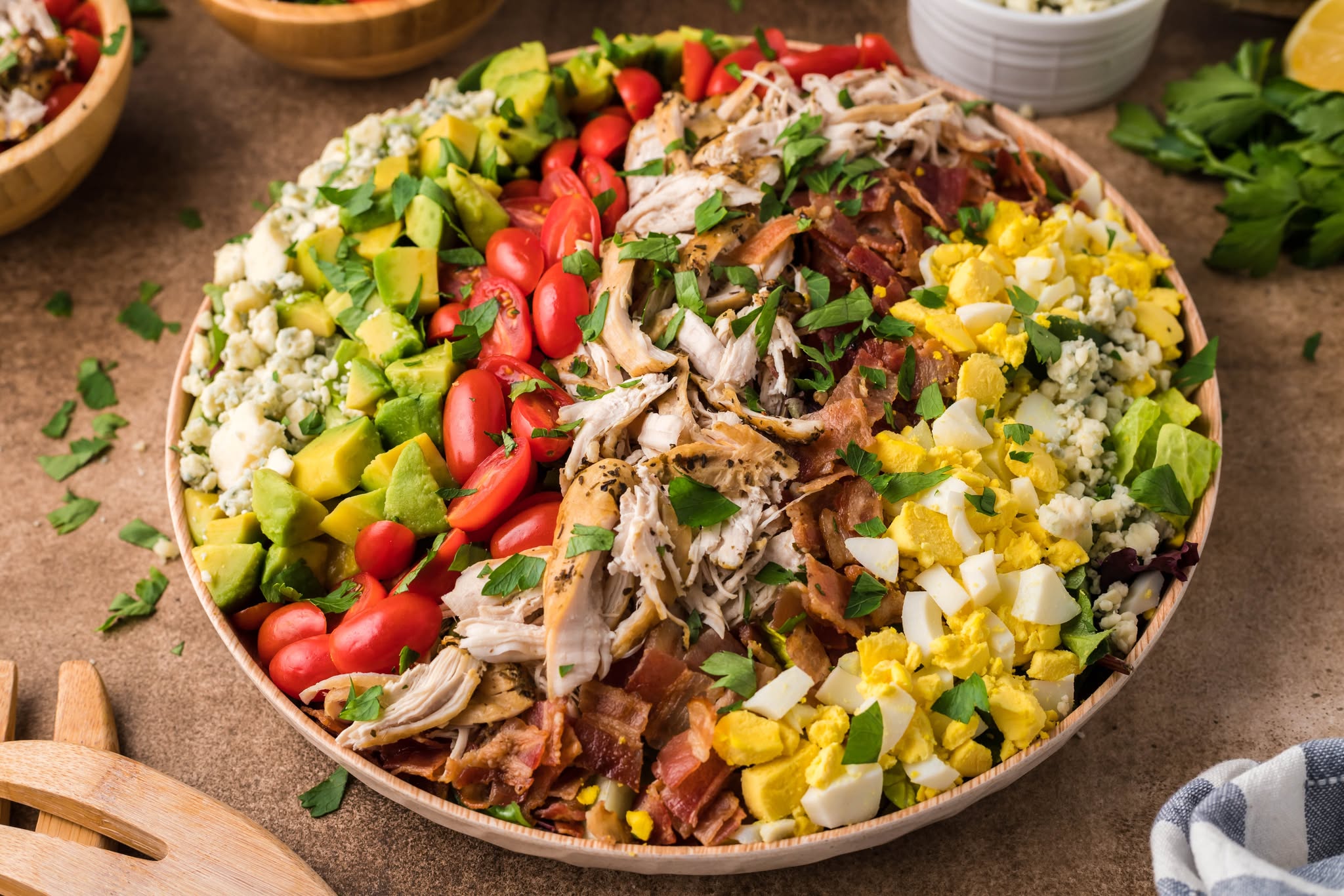 side view of the Copycat Cheesecake Factory Cobb Salad in a large wooden serving bowl on a table with serving silverware next to it