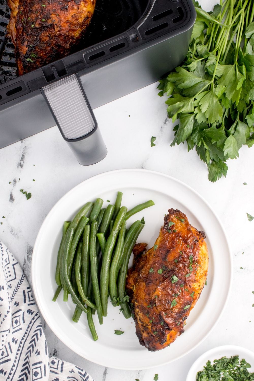 Air Fryer Chicken Breast on plate with some green beans to side and fresh herbs to side of air fryer on table