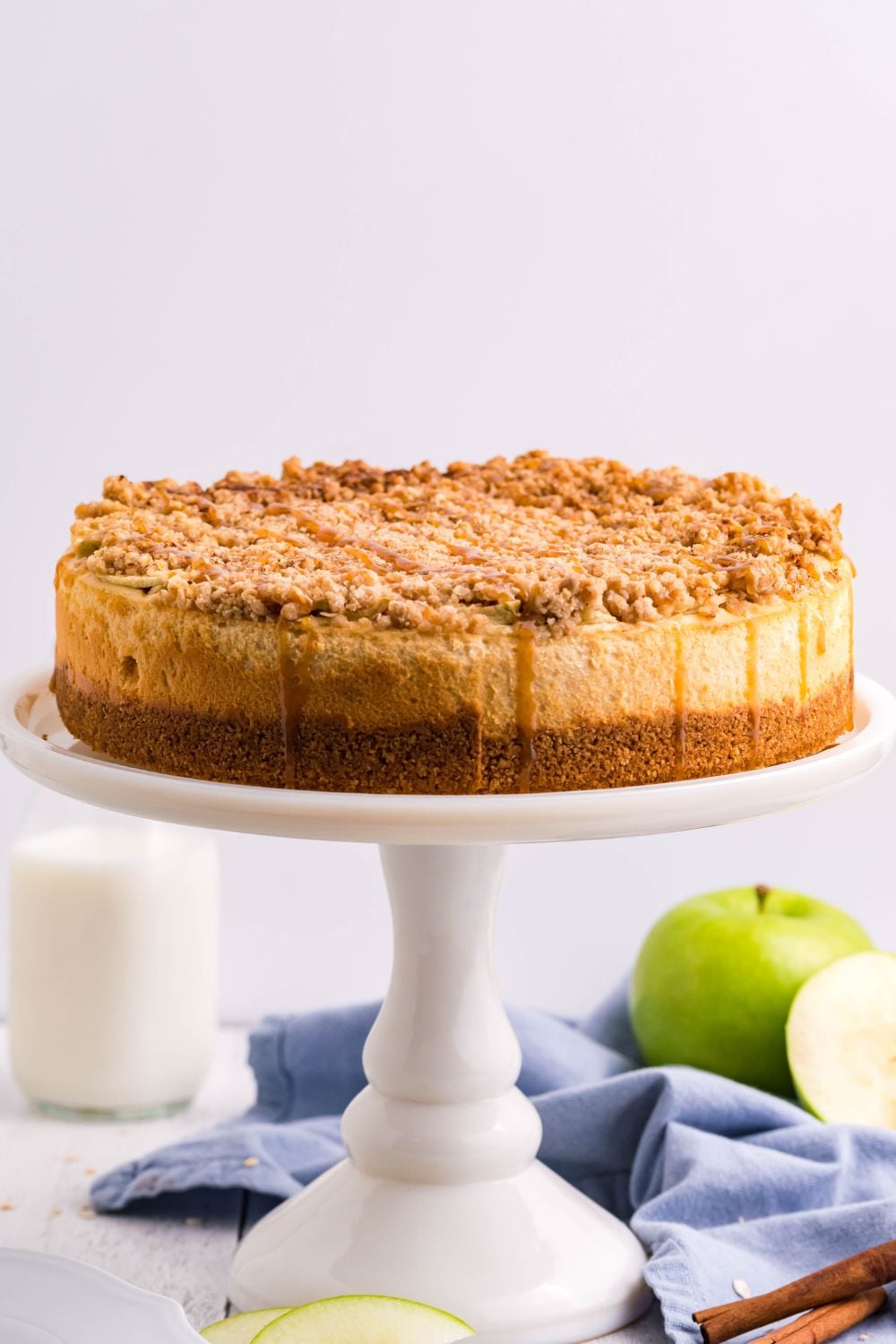 Caramel Apple Crisp Cheesecake on a cake plate with apples and milk behind it on table