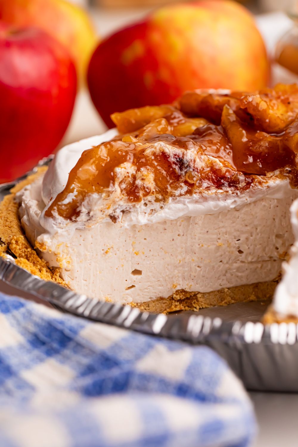 close up of No Bake Apple Cheesecake pie in pan with a slice taken out to see the layers of the cheesecake and apples in the background