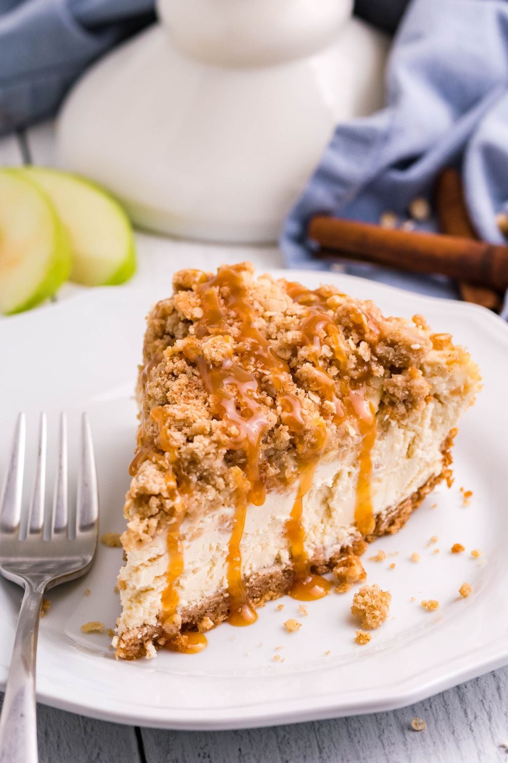 piece of Caramel Apple Crisp Cheesecake on plate iwth apples behind it and caramel sauce