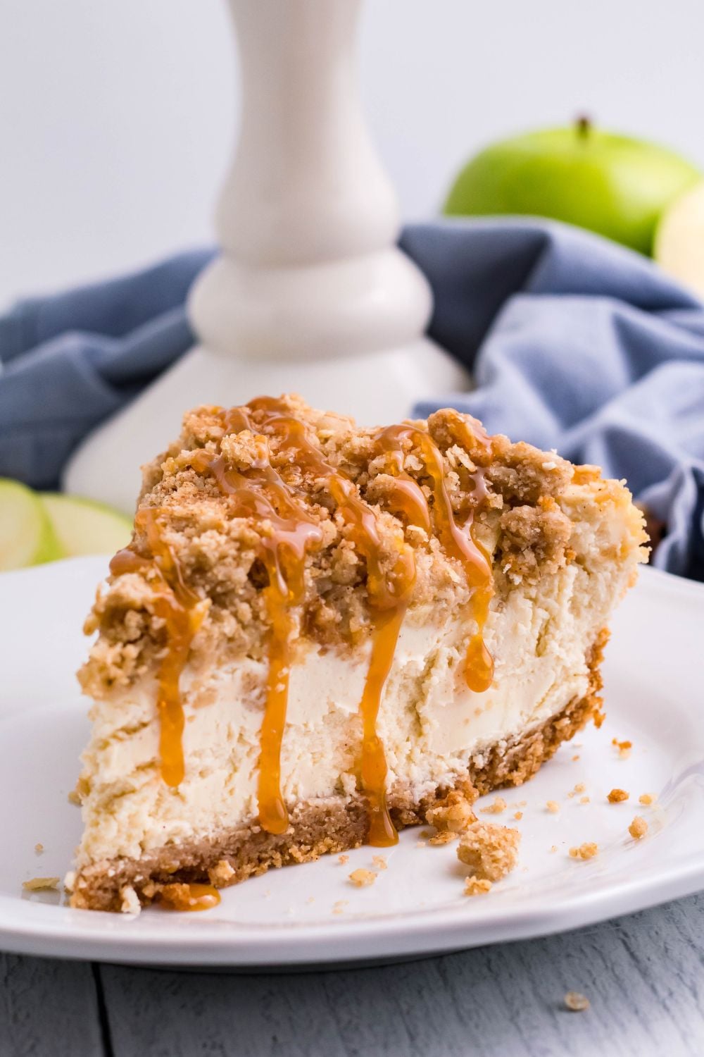 slice of Caramel Apple Crisp Cheesecake on plate with a pie plate behind it on table, napkin and green apples
