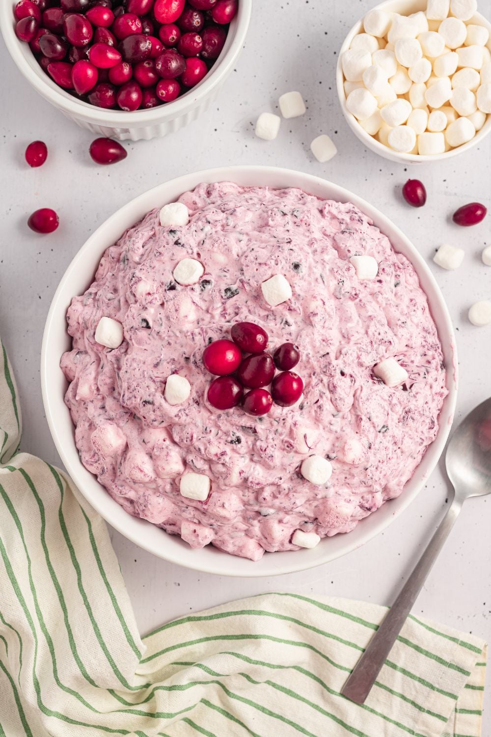 white table with a large bowl of Cranberry Fluff Salad, a spoon to side of bowl. Then you have mini marshmallows in a bowl, cranberries, and a few scattered around table