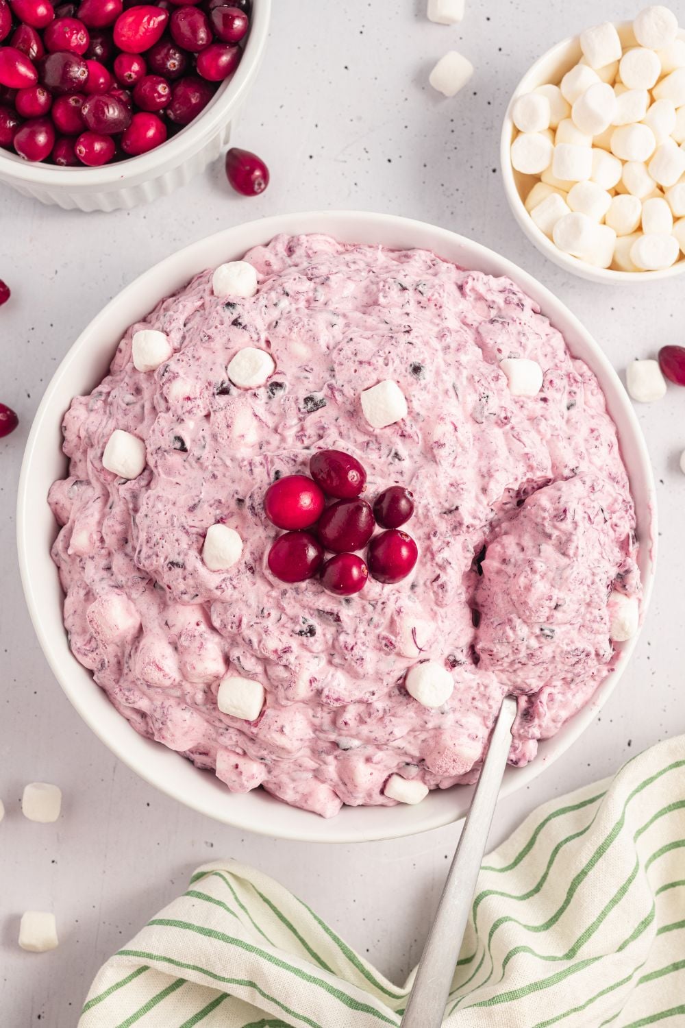 white bowl on table with some Cranberry Fluff Salad topped with extra marshmallows, cranberries, and a mini bowls of cranberries and marshmallows. Then more mini marshmallows on table with a pin striped towel to side