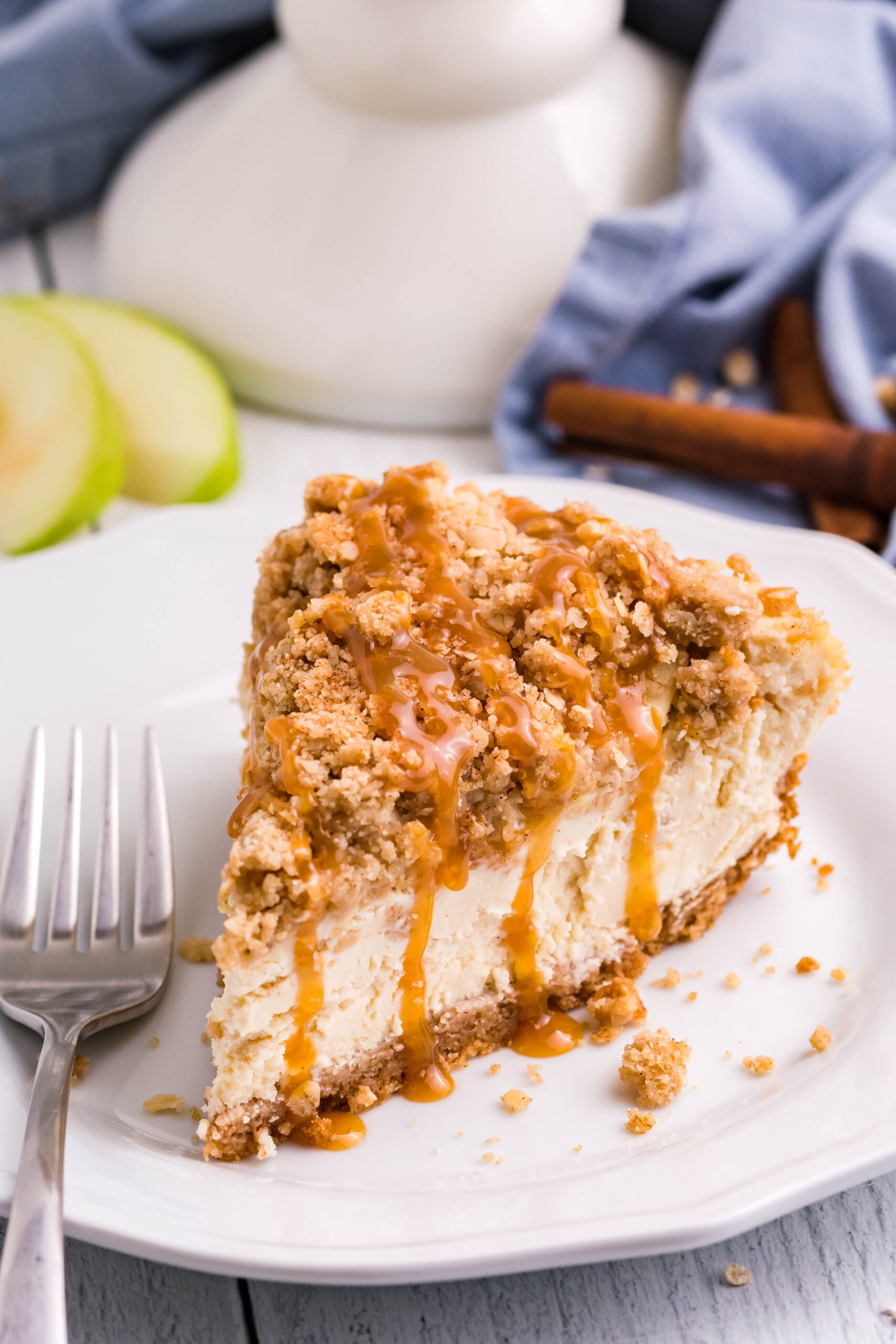 slice of Caramel Apple Crisp Cheesecake on plate with a pie plate behind it on table, napkin and green apples