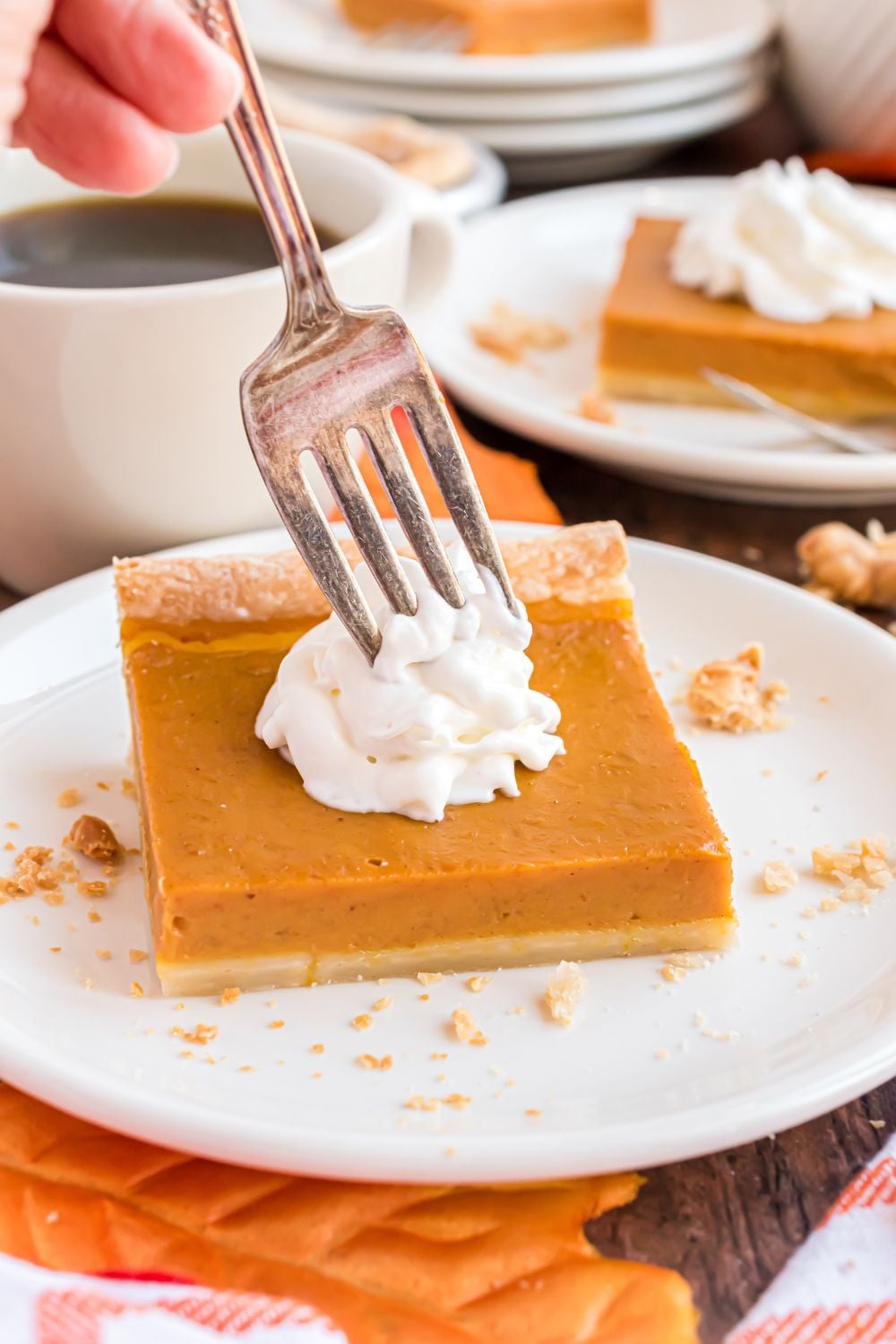 wooden table with a few white plates with slices of pumpkin pie topped with whipped cream. One plate has a fork poking into the whipped cream on top of the pie 
