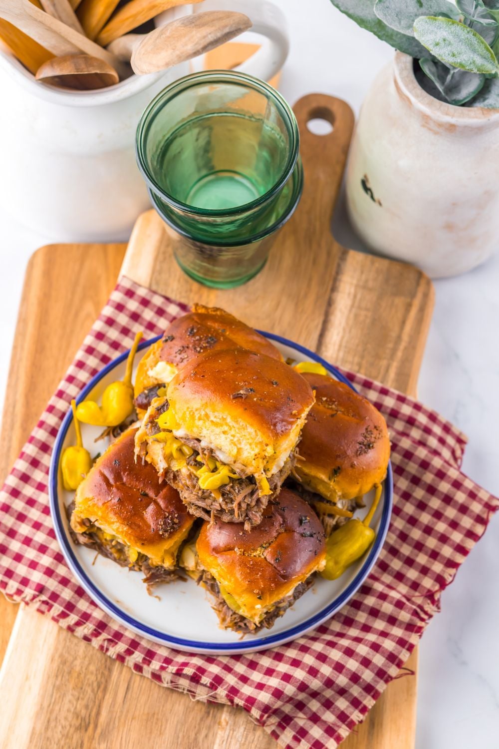 Slow Cooker Mississippi Pot Roast Sliders stacked on a plate with some glasses and ceramic containers behind it on table 