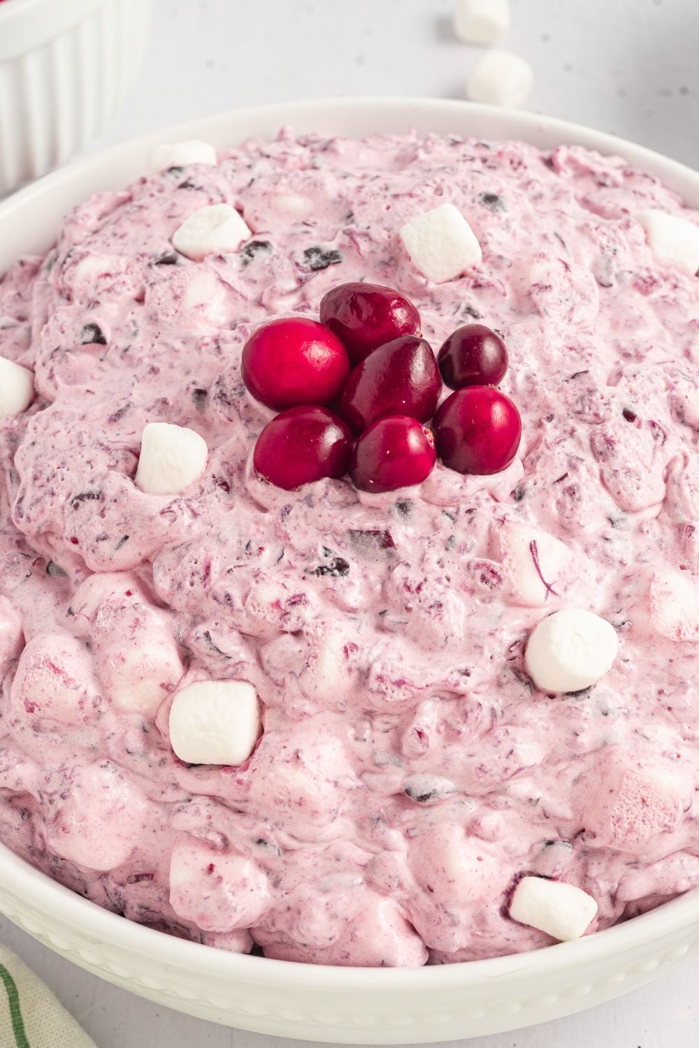 close up of a white bowl full of Cranberry Fluff Salad with mini marshmallows around the top of fluff, and in the center there are 7 fresh cranberries in the center of bowl