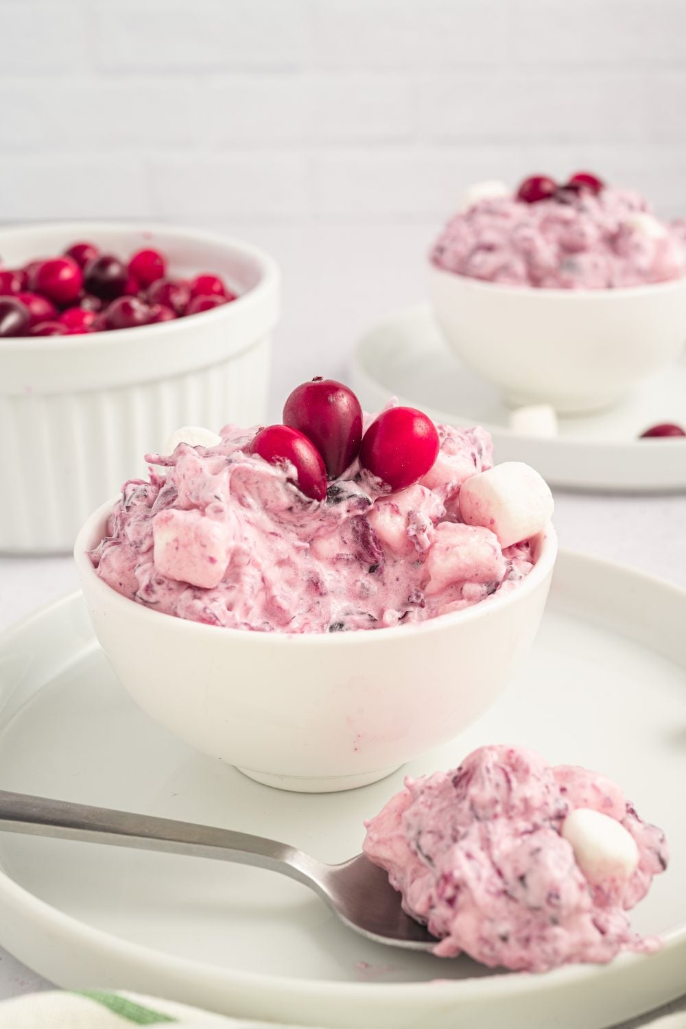 Cranberry Fluff Salad in a small bowl with a white plate under it. Then in background is another plate and bowl set with fluff inside, and a medium sized white bowl with fresh cranberries.
