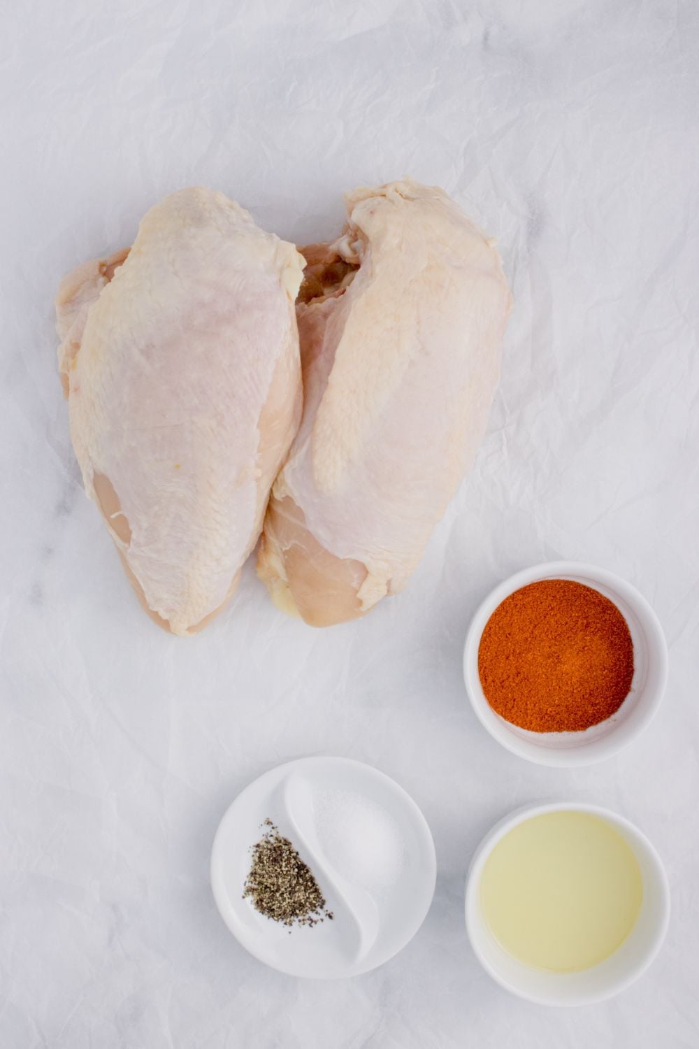 Air Fryer Chicken Breast ingredients on table, chicken breasts, seasonings in bowls, and oil in a small bowl