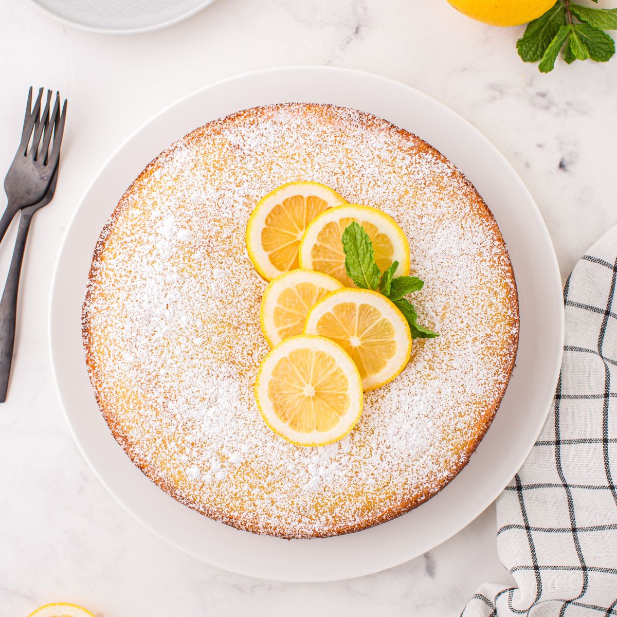 Italian Lemon Ricotta Cake on table with some fresh lemons on top with some mint leaves to side 