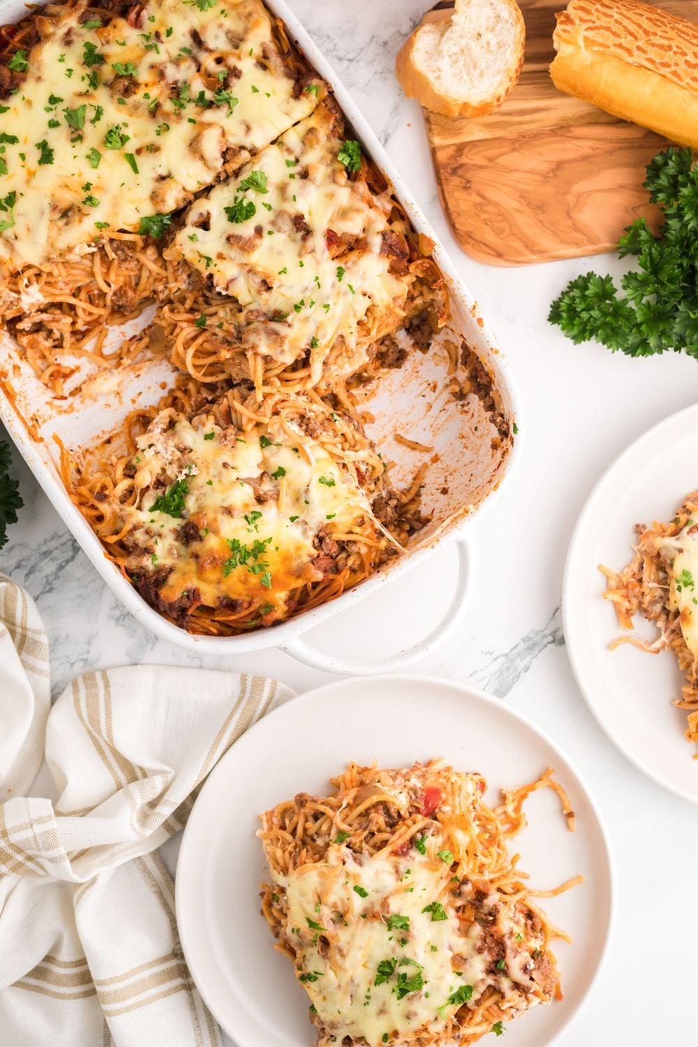 Million Dollar Spaghetti Casserole in pan with more spaghetti on plates with parsley to side and sliced bread