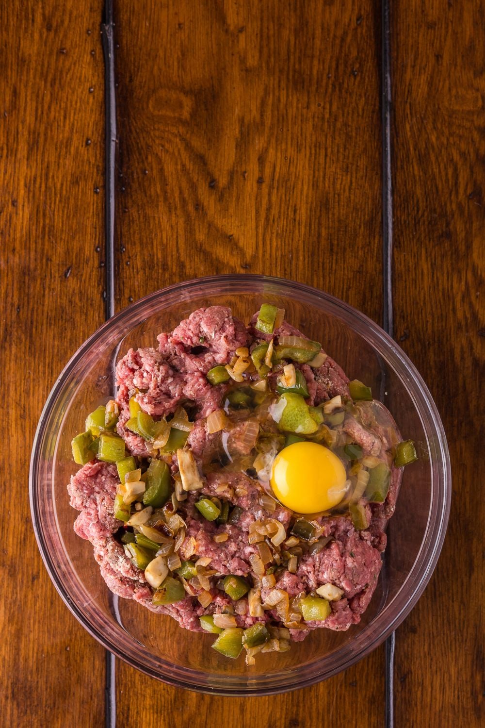 raw meat, with egg, and onions and peppers on a glass bowl on a wooden table