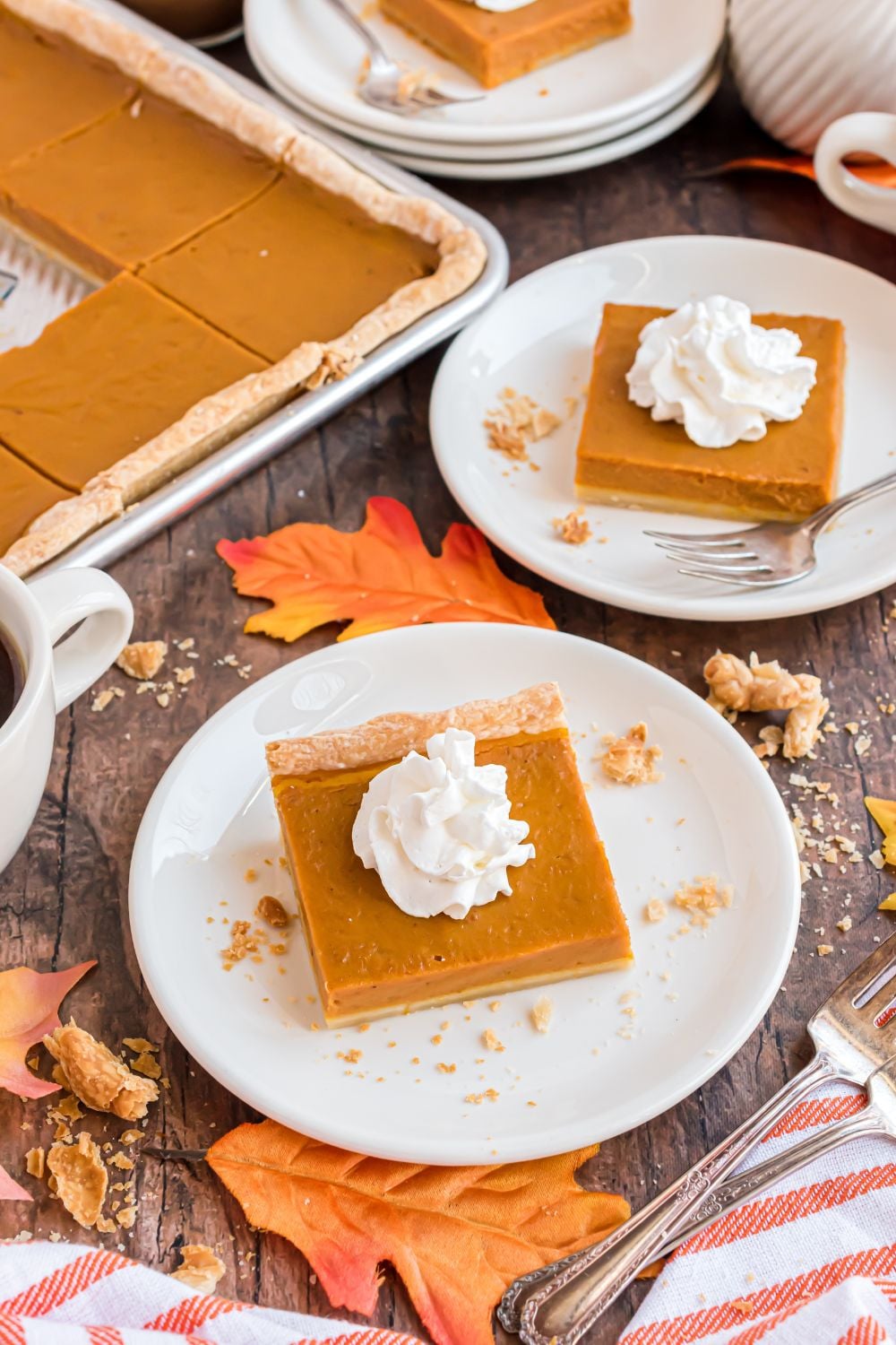 wooden table decorated for the fall season with white plates topped with slices of pumpkin pie topped with whipped cream. Then to side you have a metal sheet pan full of more sheet pan pumpkin pie 