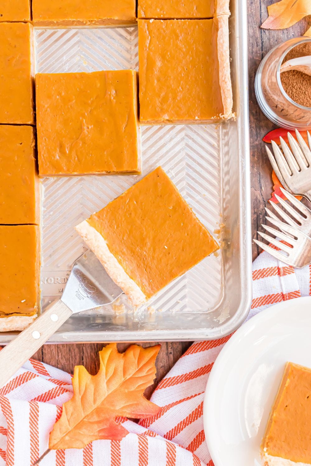 rustic wooden table with a metal sheet pan with slices of pumpkin pie in it, with a spatula lifting up more slices of pie on table 