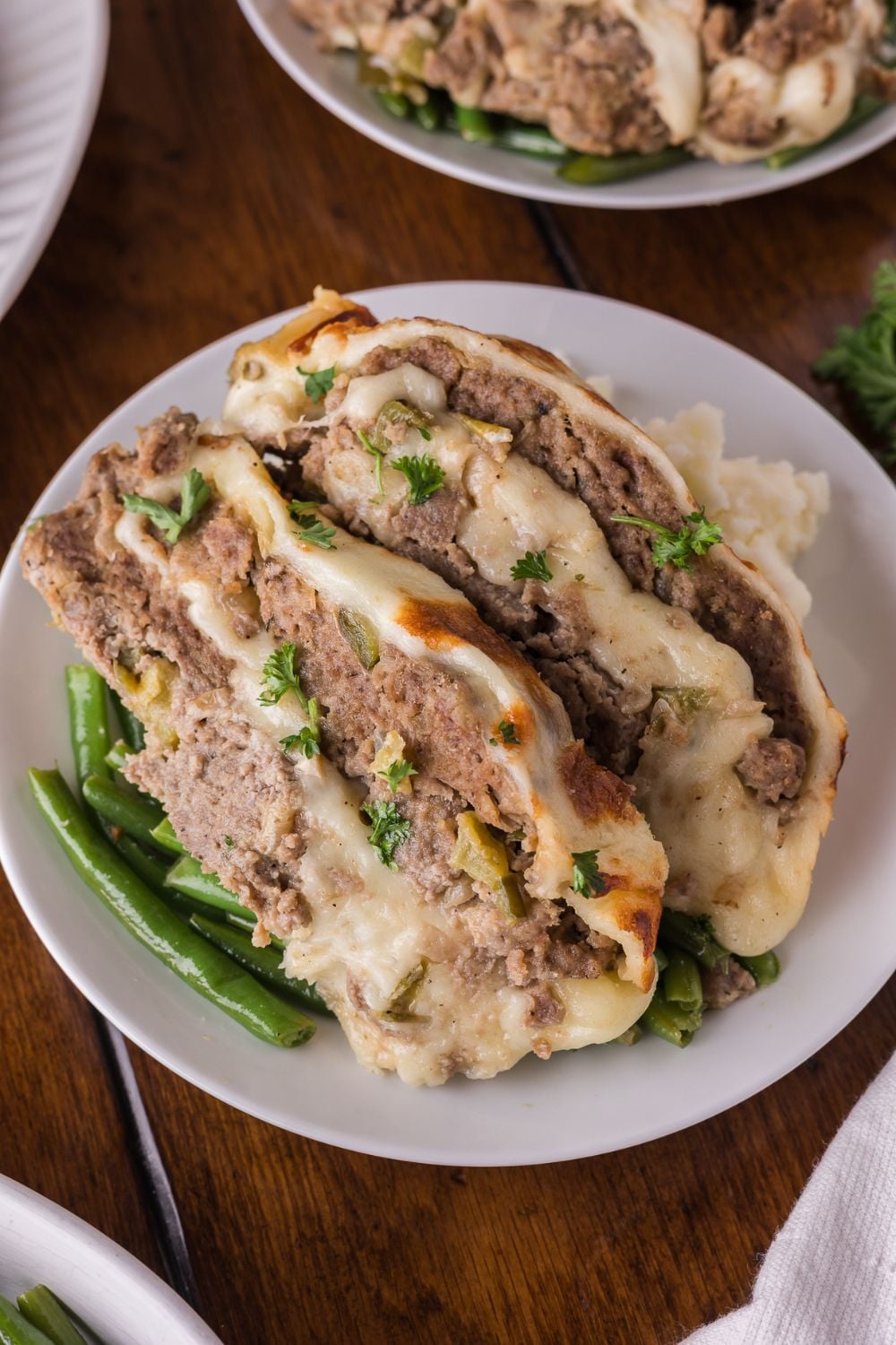 two plates on table with some green beans and large slices of Stuffed Philly Cheesesteak Meatloaf on top with mashed potatoes as well.