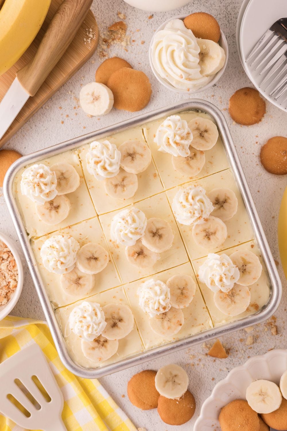 table with a pan of No Bake Banana Pudding Cheesecake Bars on it and vanilla wafers and bananas placed around the table