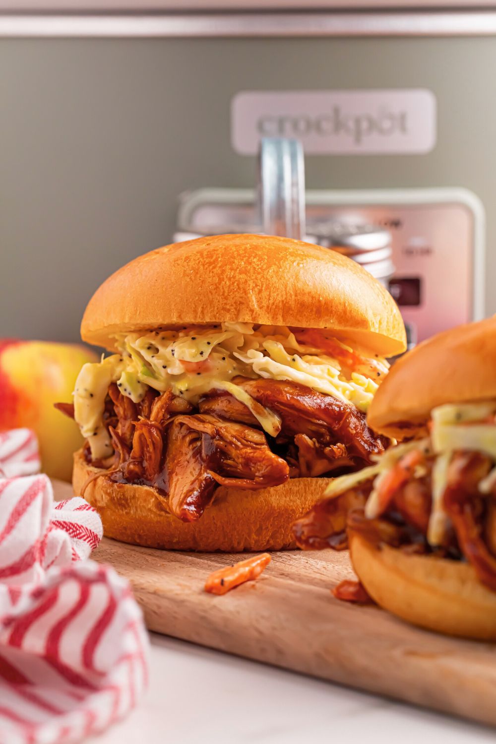 Crockpot Apple Butter BBQ Pulled Chicken sandwiches on table with a red and white towel in front and a crockpot behind sandwiches