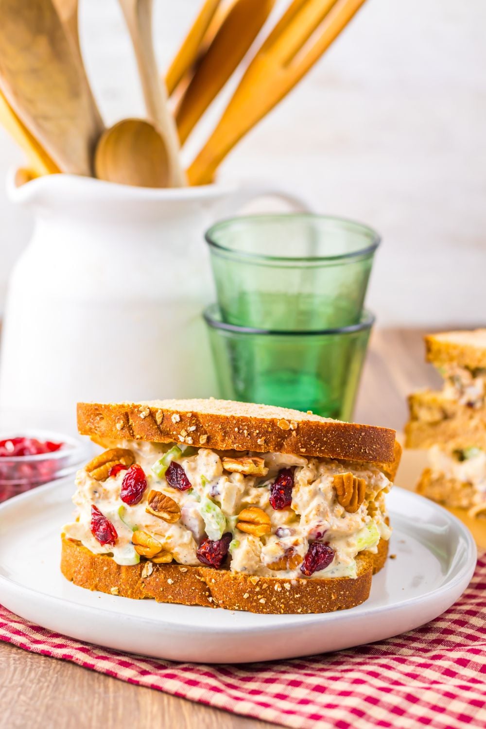 a thick turkey sandwich loaded with mayonnaise, celery, cranberries and more on a white plate on table with a checkered napkin spread out under the plate, with a rustic look to the table 