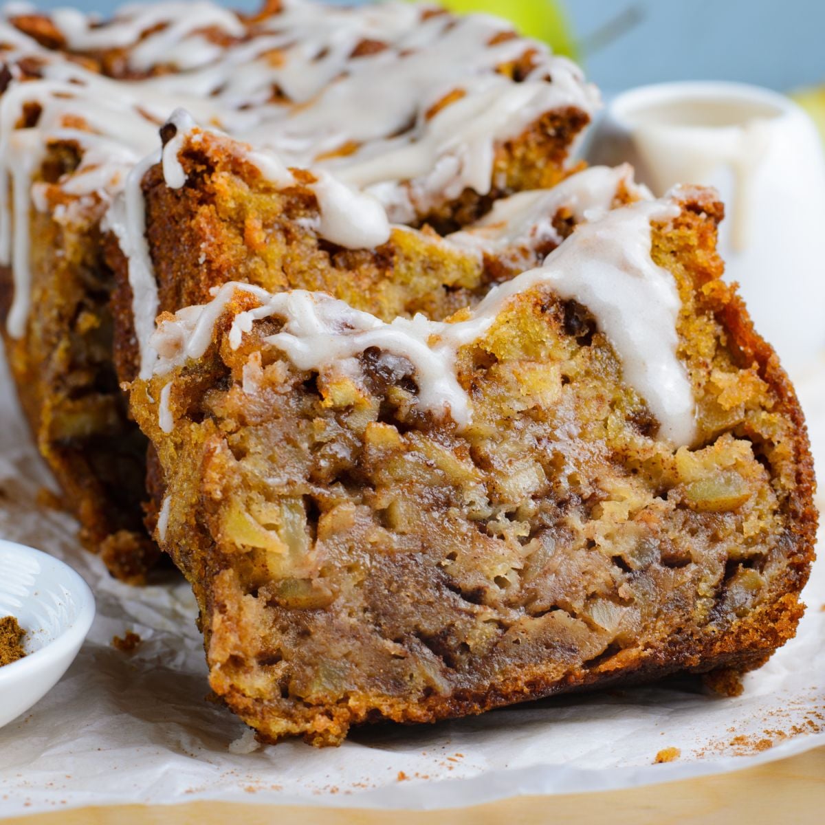 Amish Apple Fritter Bread sliced and placed on a parchment paper on table with a bowl with glaze to the side