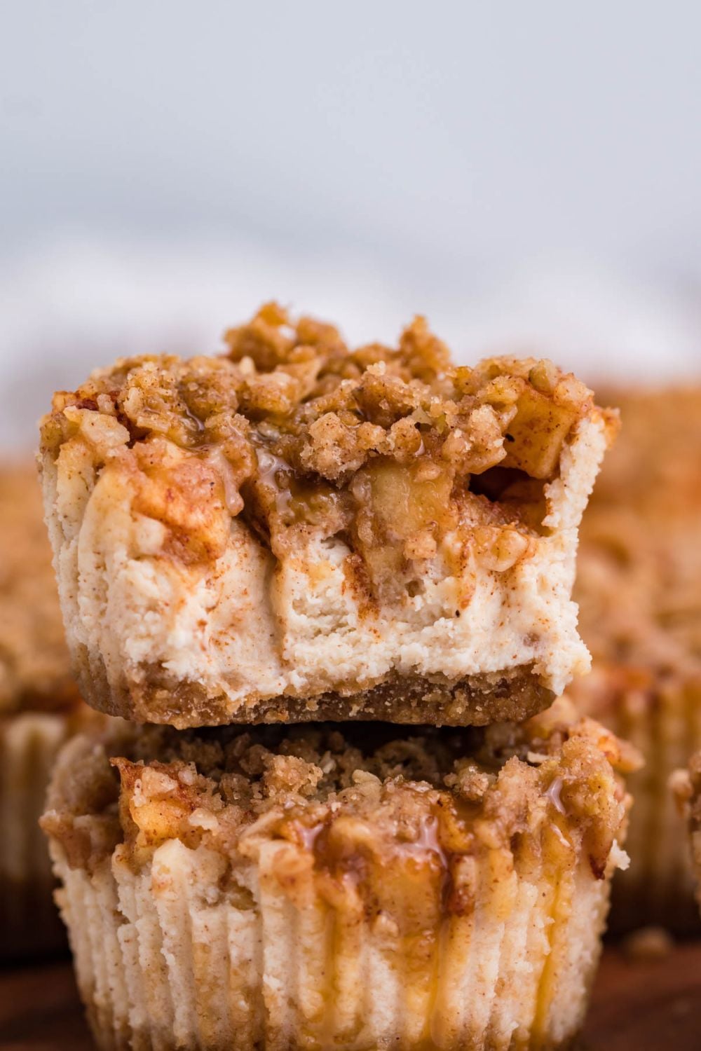 two Apple Crisp Cheesecake Bites stacked on top of each other on wooden cutting board. The top cheesecake has a few bites taken out of it