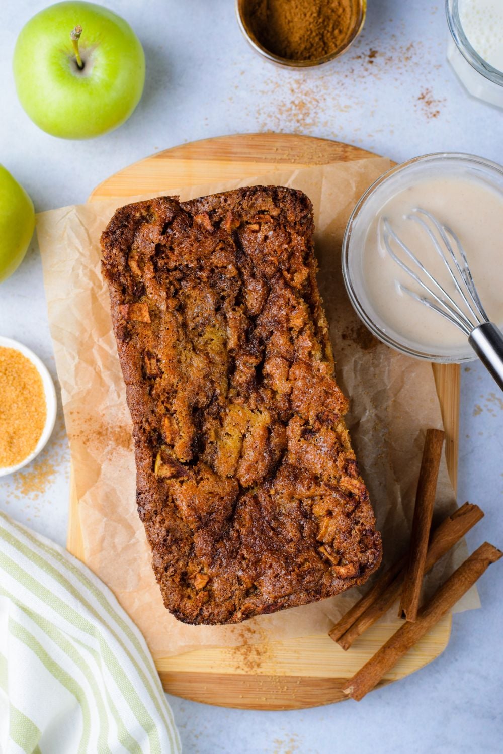 apple fritter bread sitting on marble counter top with glaze in a bowl to side, cinnamon sticks, apples and more scattered around table