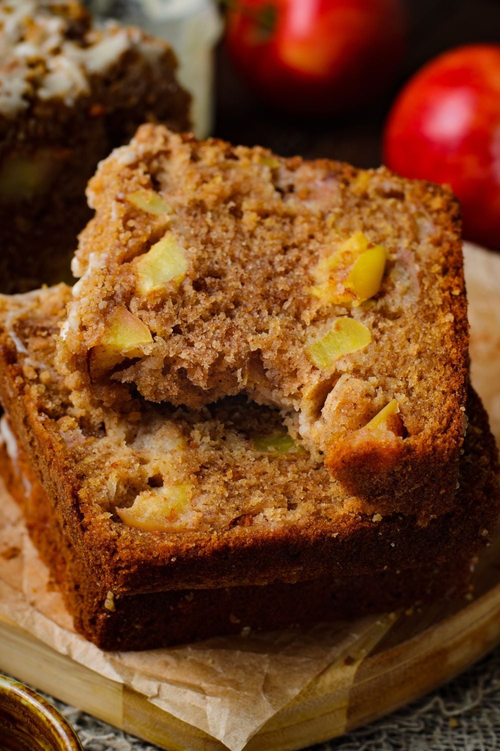 Apple Cinnamon Loaf Cake sliced into thick pieces stacked on top of one another on a tray. The top slice has a bite taken out, and red apples are behind it on table for a nice red color to image