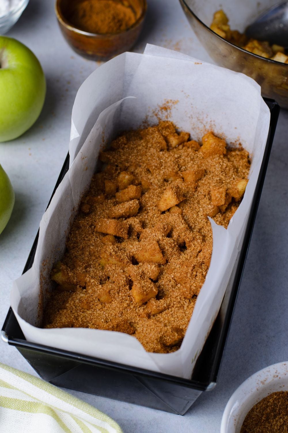 cinnamon mixture and diced apples inside a loaf pan sitting on marble table with green apples to side of pan