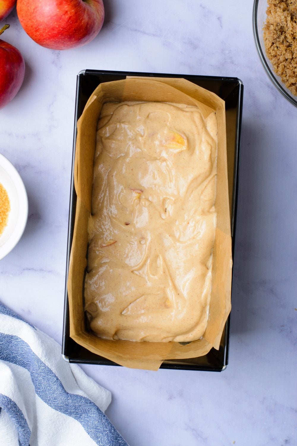 looking down at marble counter with a loaf pan with half full of a cinnamon loaf cake batter and apples and a brown sugar mixture to the side