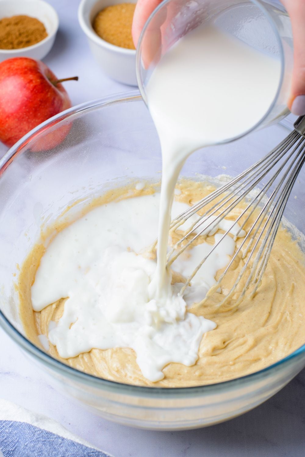 wet batter in a glass bowl with some milk pouring in and a whisk to the side. A red apple is on the table as well