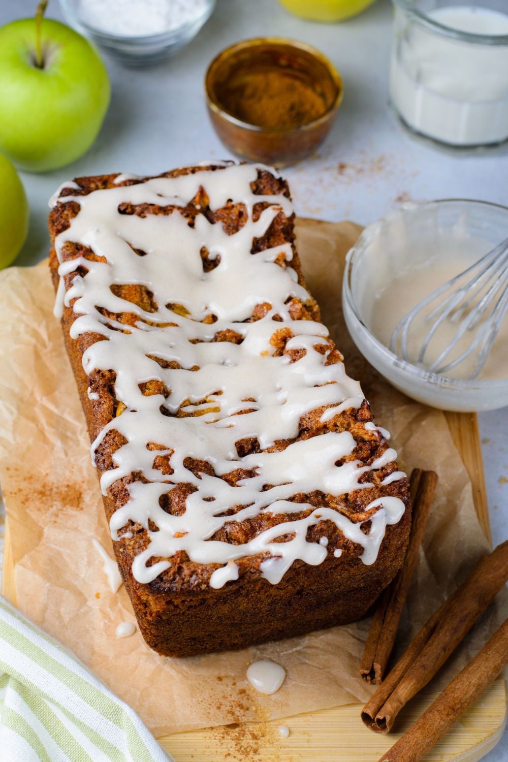 Amish Apple Fritter Bread on brown parchment paper on table with a bowl of glaze to side and cinnamon sticks in front of loaf