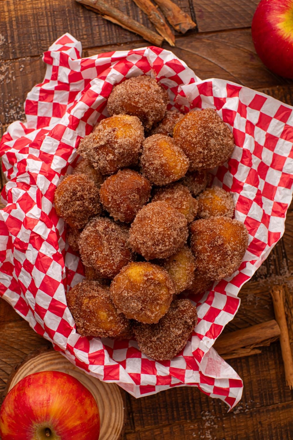 looking down on a wooden table with a basket full of Apple Pie Donut Holes and whole apples and cinnamon sticks around table 