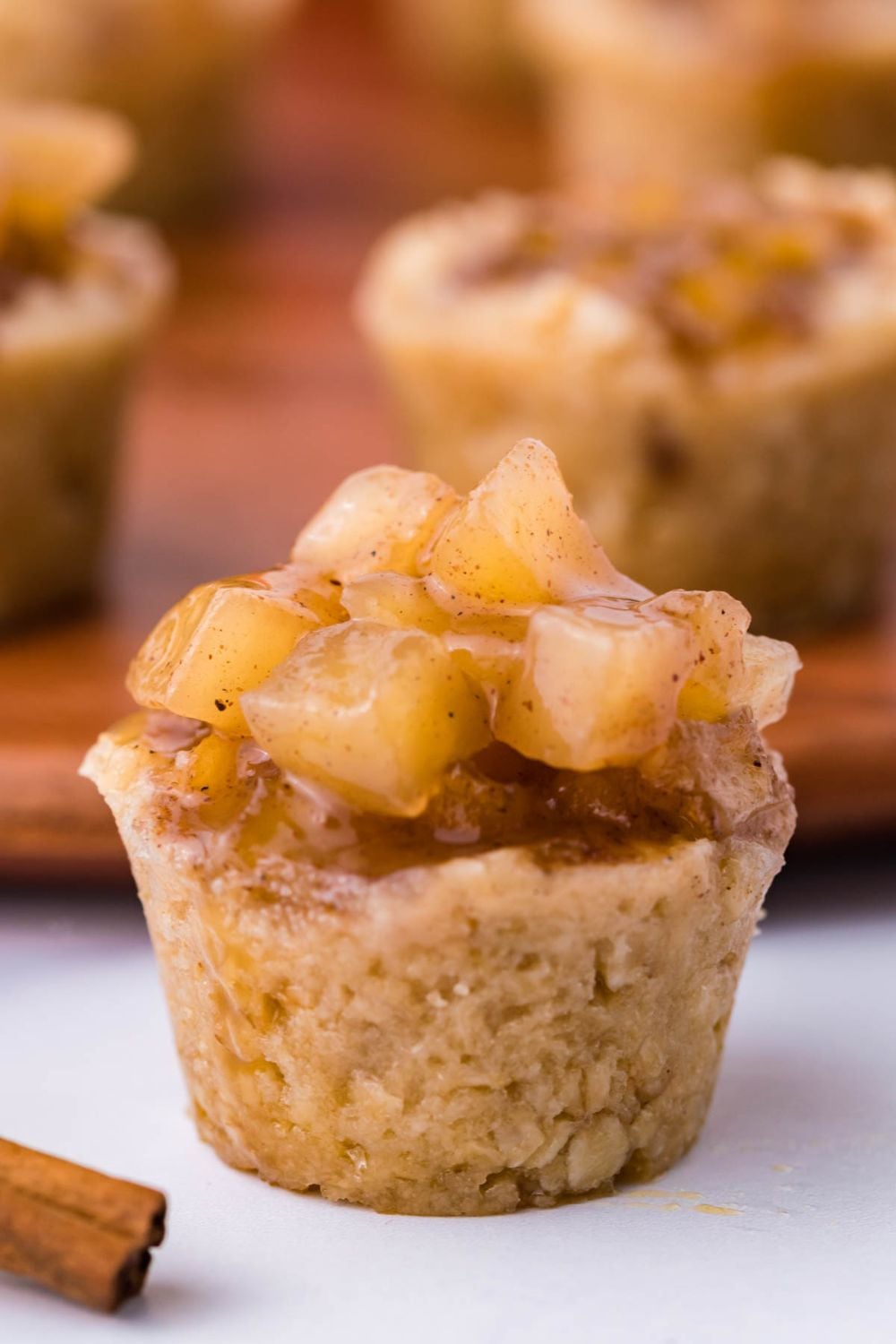 close up look of a Mini Apple Crisp Bites on white table with a cinnamon stick in front, and more bite cups behind it on a wooden tray
