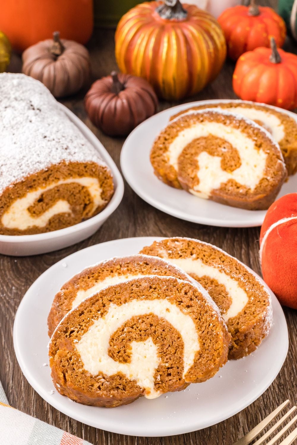 Pumpkin Cake Roll placed on plates, with a small platter to side with more of the pumpkin roll. Mini pumpkins scattered on table for a Thanksgiving fall look