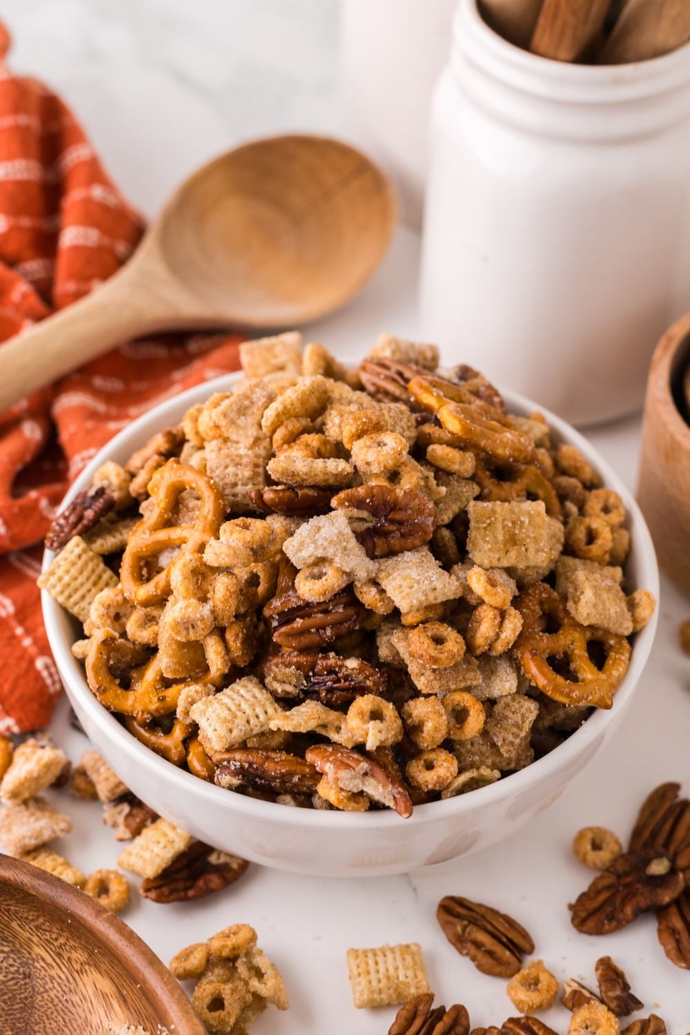orange towel laid out on table with a wooden spoon in the back, and a bowl full of Pumpkin Spice Chex Mix and more snack mix spread around the table 