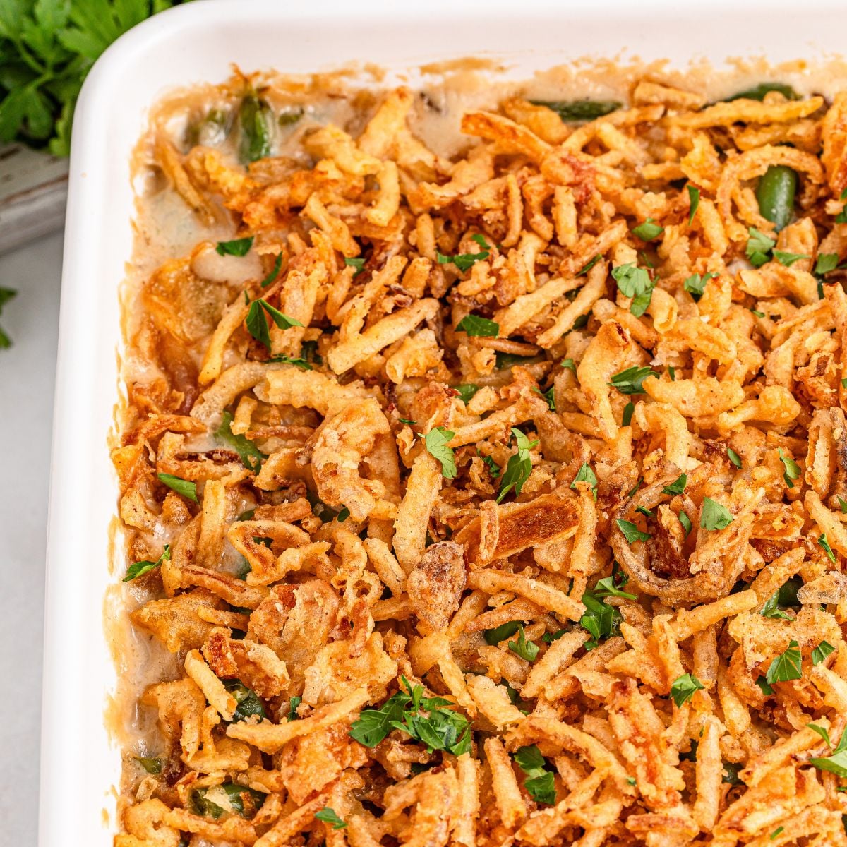Green Bean Casserole in a white casserole dish sitting on table with fresh parsley sprinkled on top of casserole 