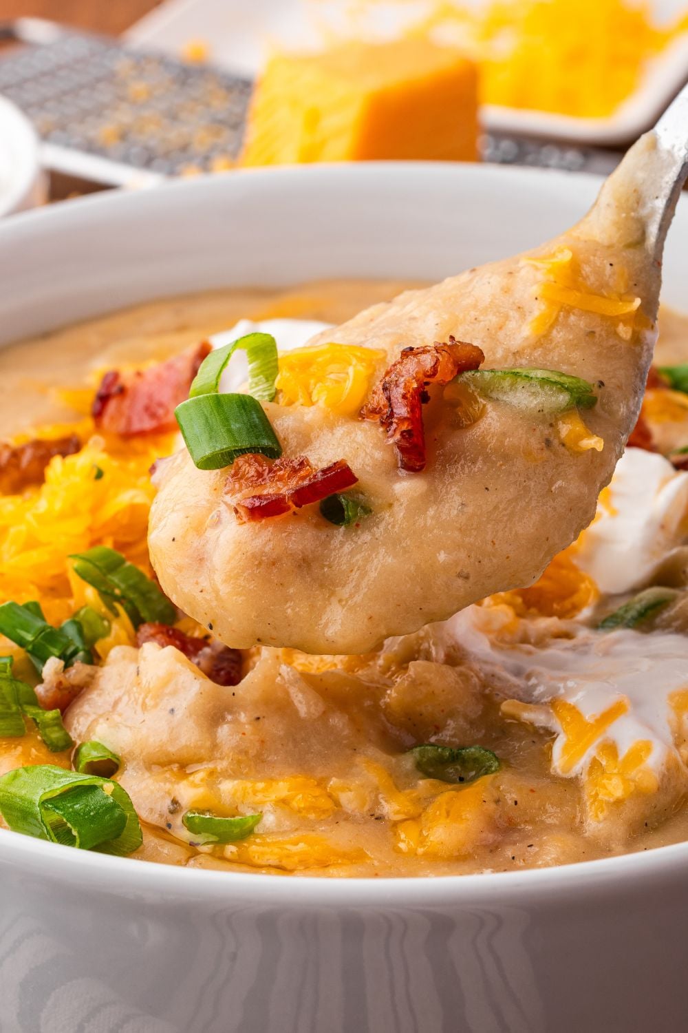 close up of white bowl on table with Crockpot Loaded Baked Potato Soup in it, and topped with crumbled bacon, green onion and shredded cheese. In the background there is a cheese grater with a block of cheese