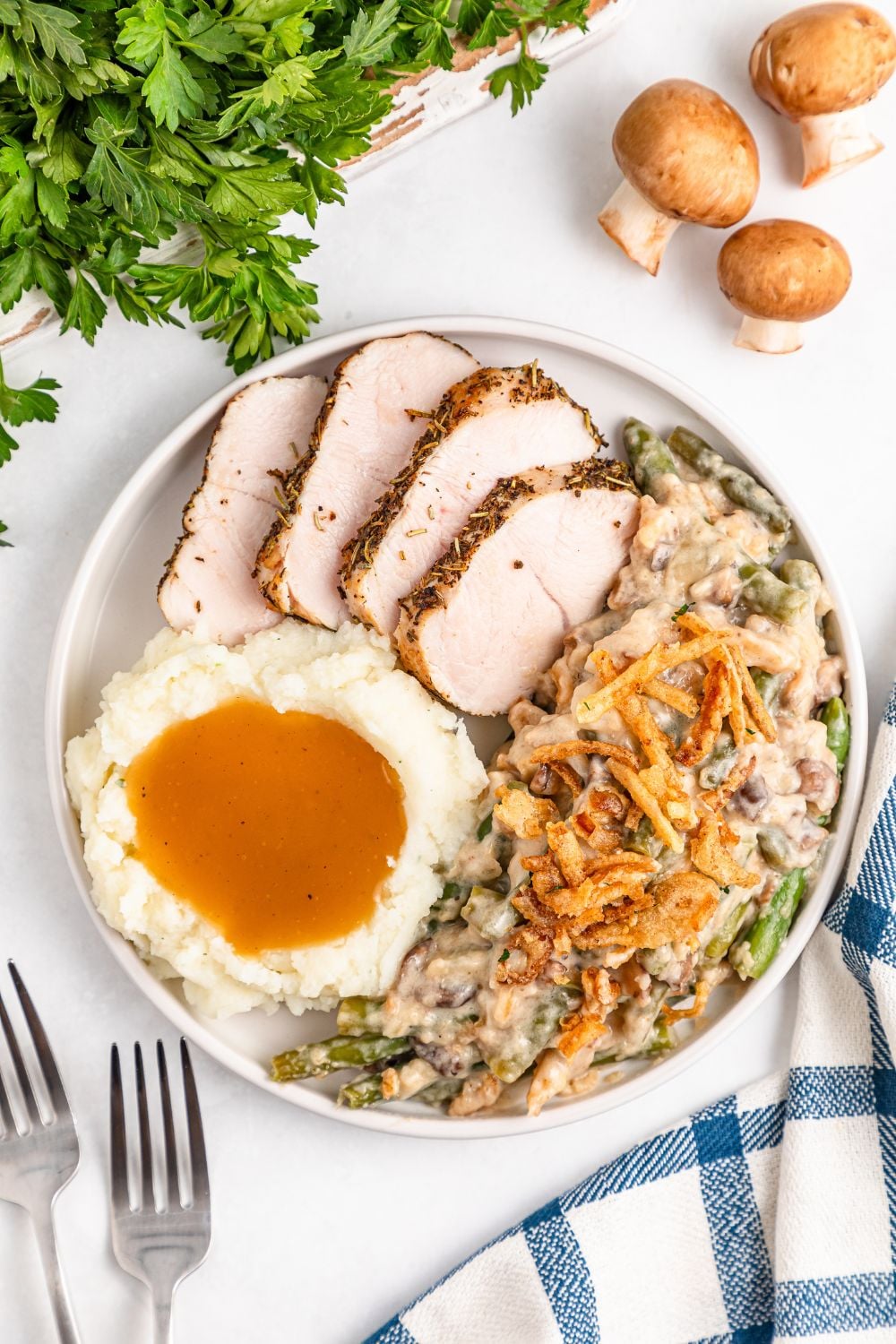 looking down on a white table with a plate full of holiday food- sliced turkey, mashed potatoes, gravy, and green bean casserole. Mushrooms and parsley are around table for decor 