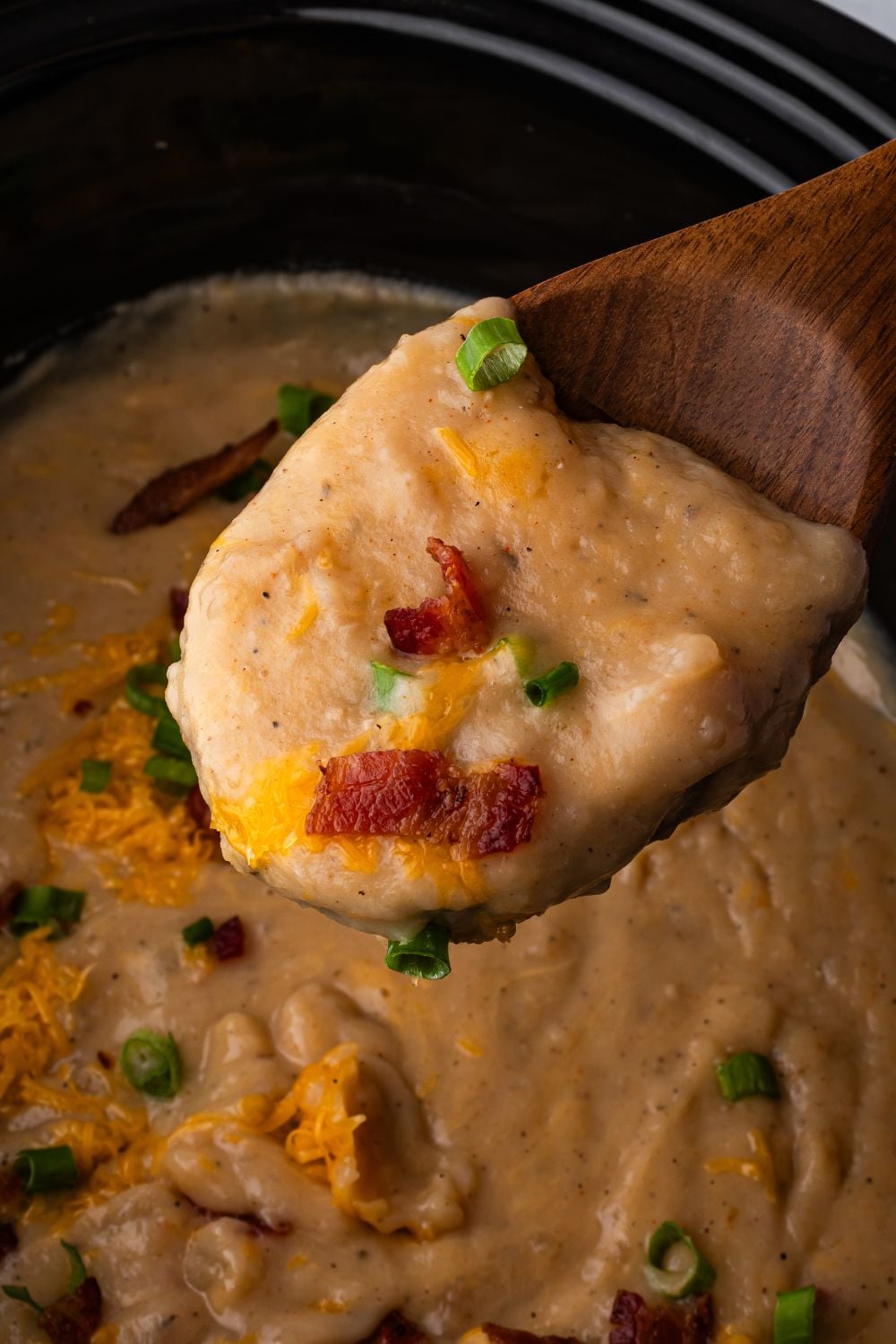 wooden bowl with some of the loaded potato soup with cheese, bacon and green onions in a black crockpot