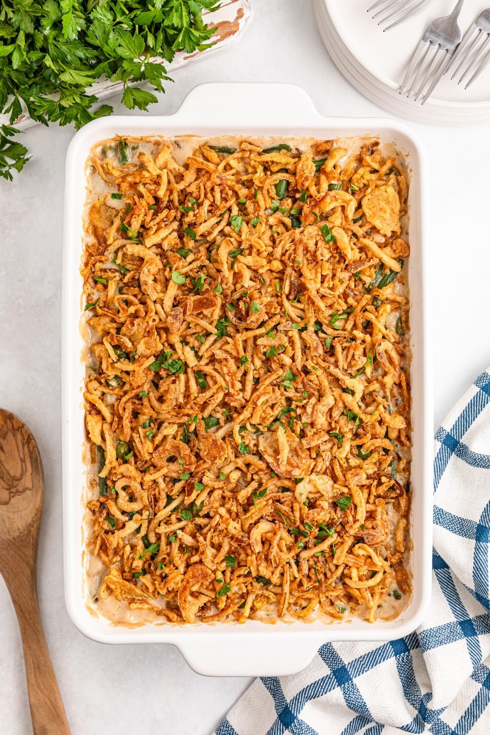 Green Bean Casserole in a white dish on table with a wooden spoon, towel and more placed around pan 