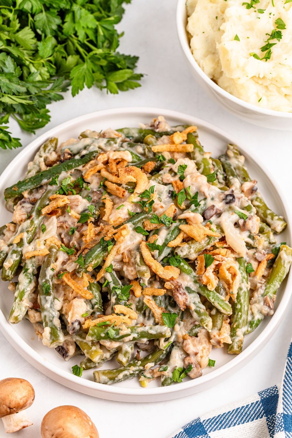Christmas green bean casserole in a round white dish on table with mashed potatoes in a bowl to side and fresh herbs
