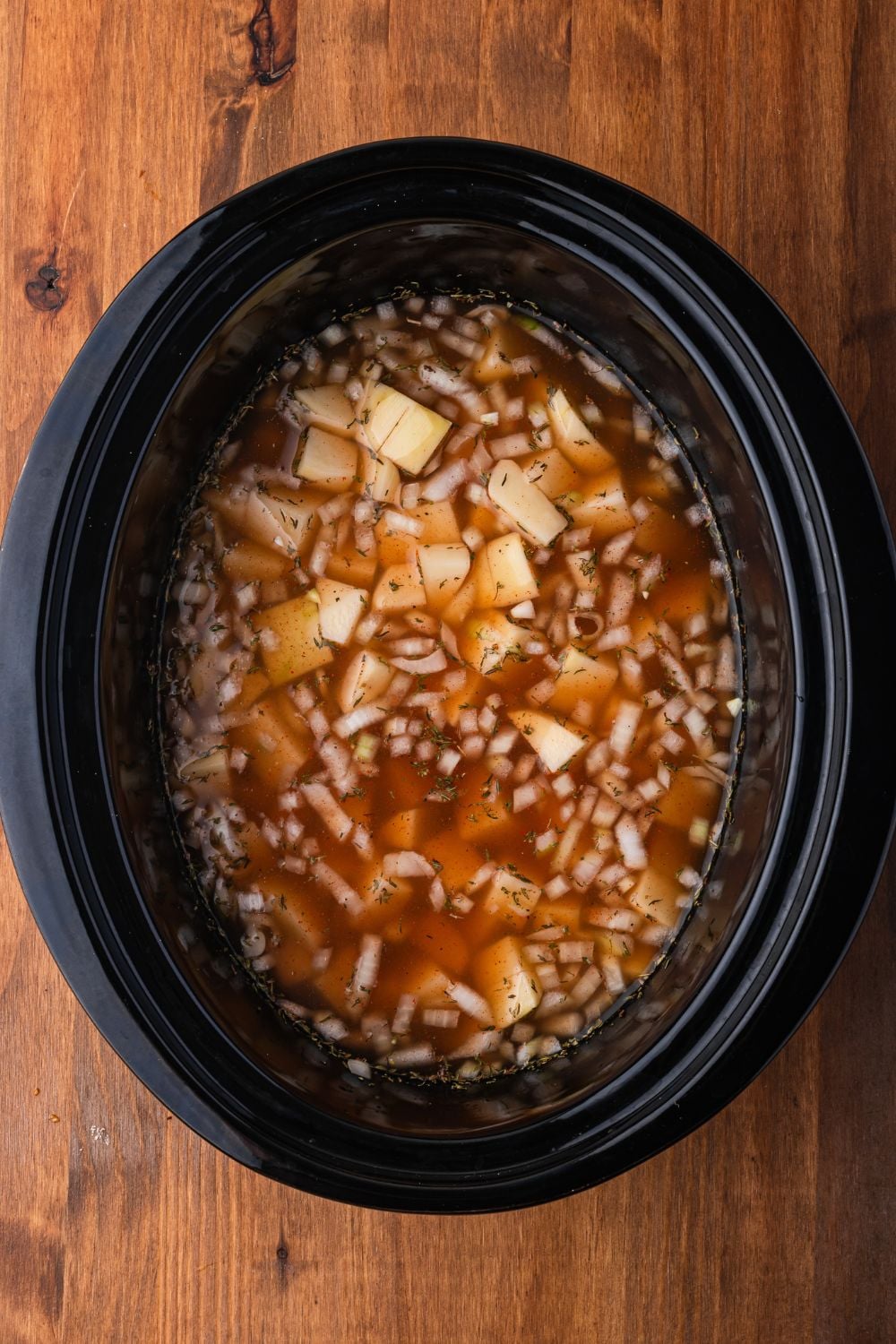 black crockpot sitting on a wooden table with diced potatoes, onions, beef broth in more in the slow cooker