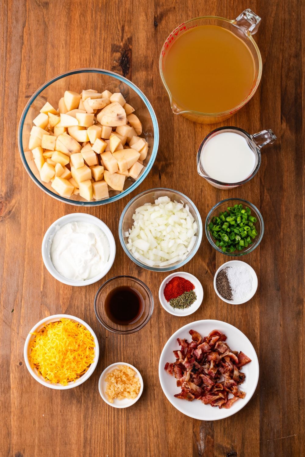 wooden table with all the ingredients in single serve bowls for making Crockpot Loaded Baked Potato Soup. Potatoes, bacon, onions, cheese, and all the other ingredients.
