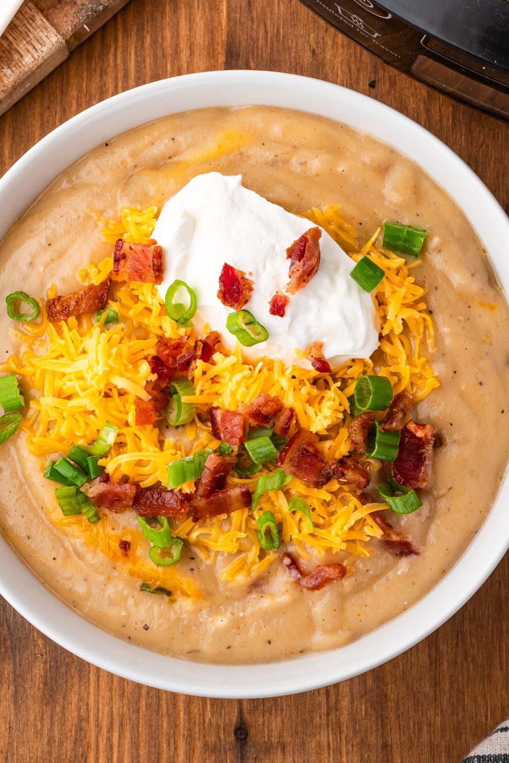 looking down at table with a white bowl full of Crockpot Loaded Baked Potato Soup and topped with crumbled bacons, shredded cheese, sour cream and more.