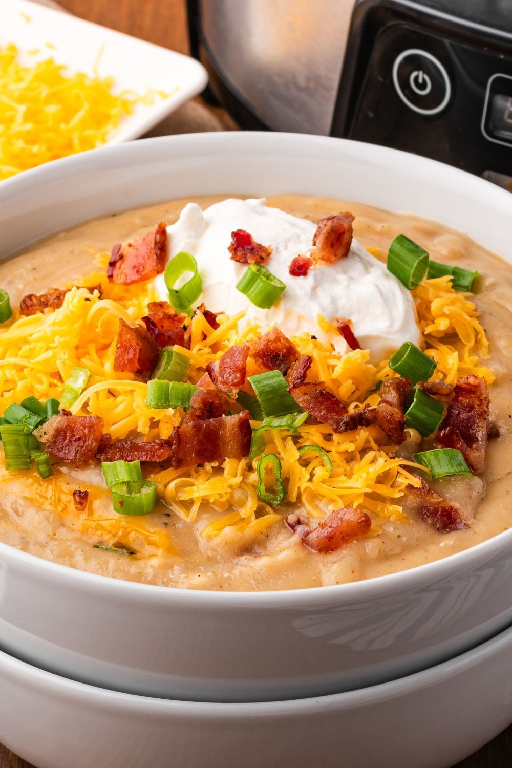 two white bowls stacked and top one full of Crockpot Loaded Baked Potato Soup with toppings . Behind the bowls is a silver and black crockpot and a small bowl of shredded cheese.