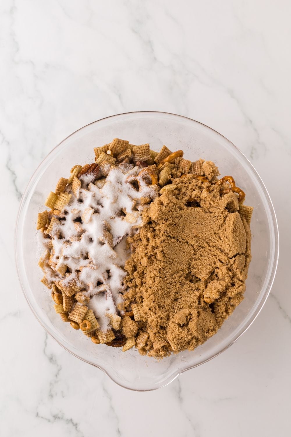 glass bowl on marble counter with Pumpkin Spice Chex Mix ingredients inside topped with brown sugar 